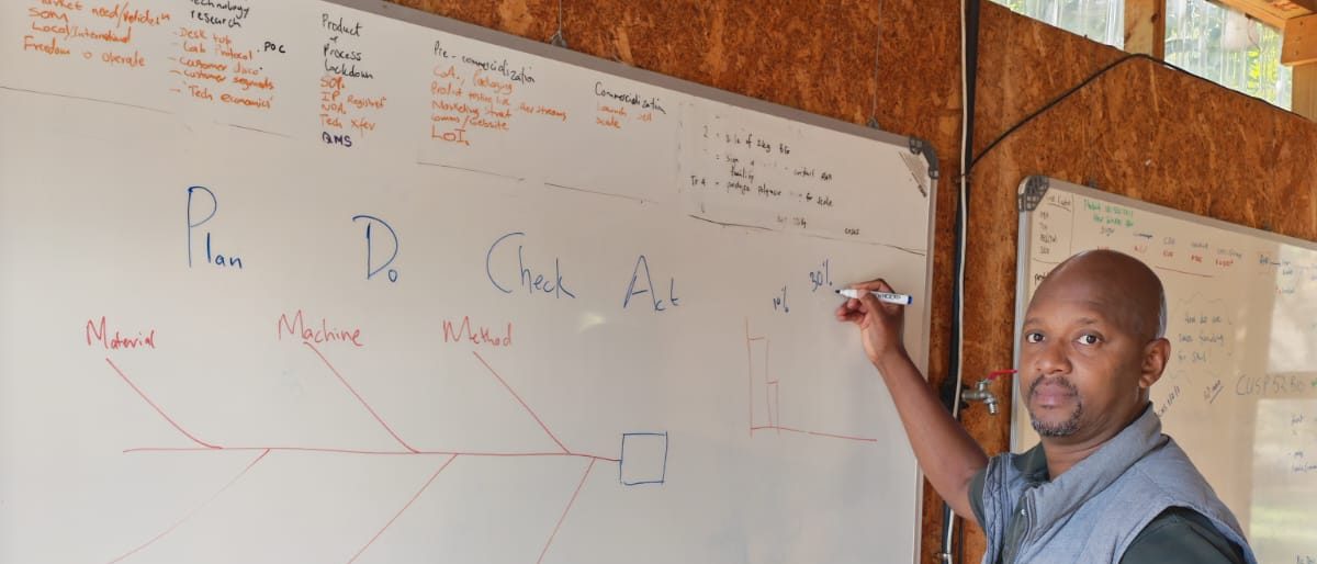 An SMP consultant at the whiteboard running a structured problem-solving session — the PDCA cycle (Plan, Do, Check, Act) and an Ishikawa root-cause diagram mapped out with a production team